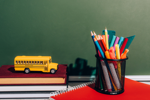 toy school bus on a stack of books, cup of pens on a red notebook